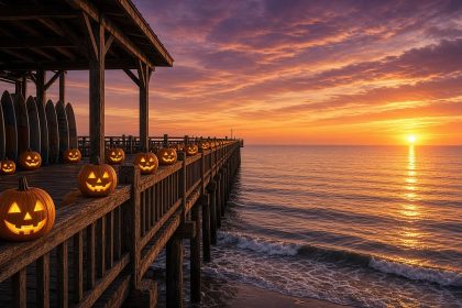 Glowing jack-o'-lanterns line the railing of a wooden beach-town pier at sunset, with surfboards stacked nearby and the Pacific glowing orange and purple.